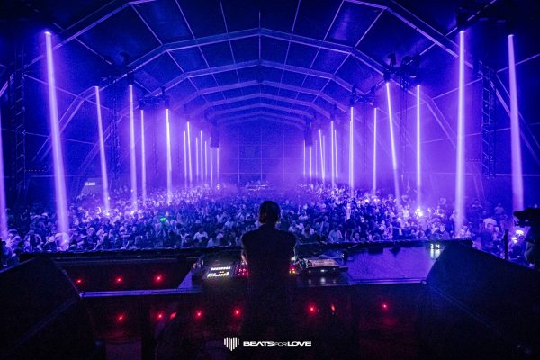 DJ performing in front of a large crowd under a tent with vertical purple lights illuminating the scene at Beats for Love festival
