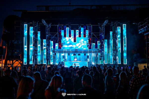 Crowd watching a DJ performing on stage with vertical blue LED light panels at the Beats for Love music festival at night.