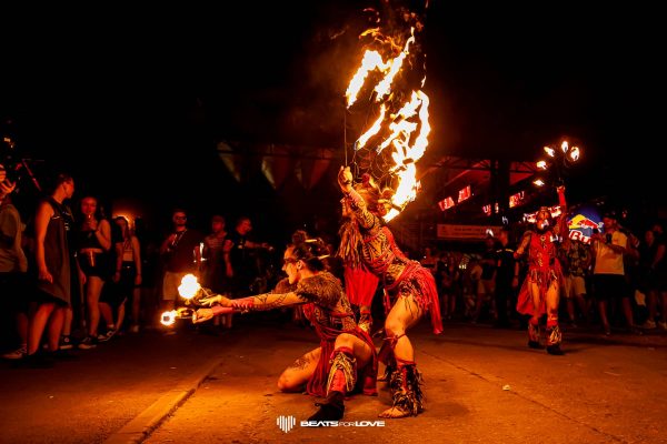 Two fire dancers in tribal costumes performing with flaming props in front of a nighttime crowd at an outdoor event.