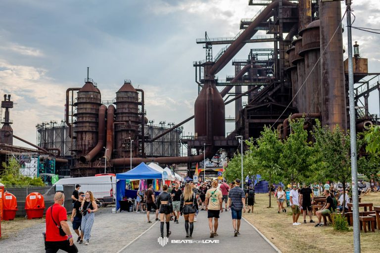 Crowd walking along a festival path with food stalls and old industrial structures in the background at Beats for Love event.