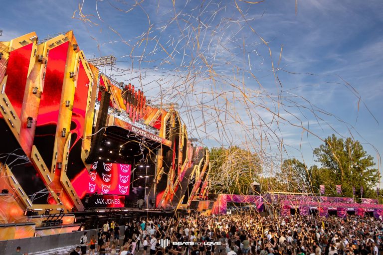 Outdoor music festival stage with red and orange geometric designs, crowd of people, and confetti streams in the air.