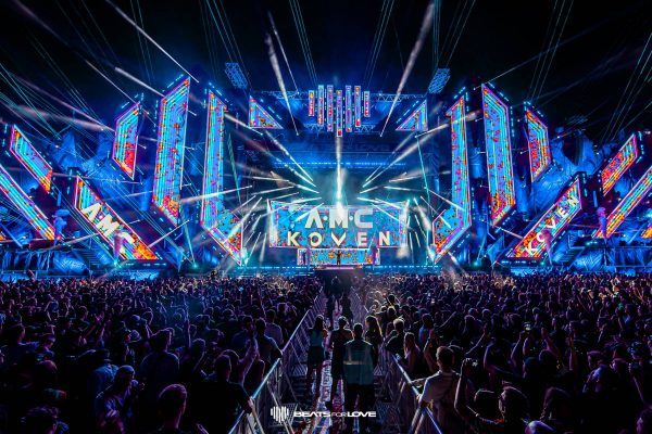 Large crowd at a nighttime electronic music festival with bright blue and red LED stage lights displaying 