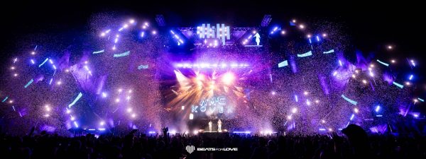 Large outdoor music festival stage with bright purple and blue lights, confetti, and a crowd silhouetted in front.