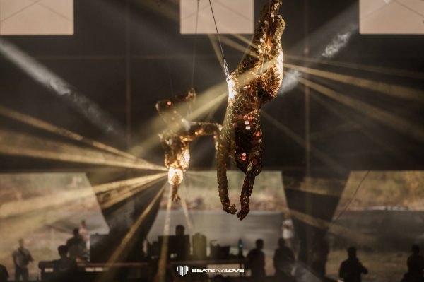 Two gold honeycomb-textured suits suspended in air with spotlight beams cutting through fog and silhouettes of people below