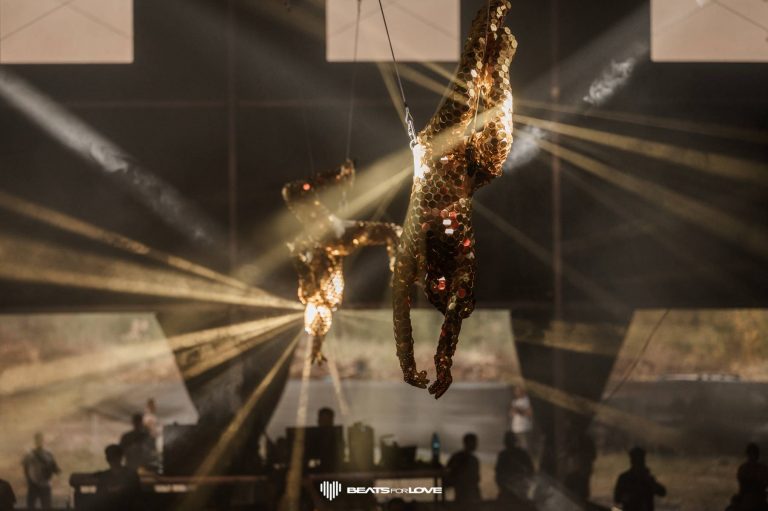 Two gold honeycomb-textured suits suspended in air with spotlight beams cutting through fog and silhouettes of people below