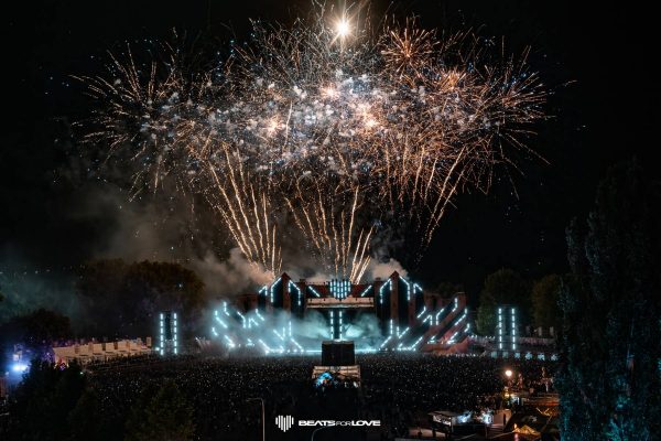 Nighttime music festival with a large crowd, illuminated stage, and golden fireworks exploding overhead