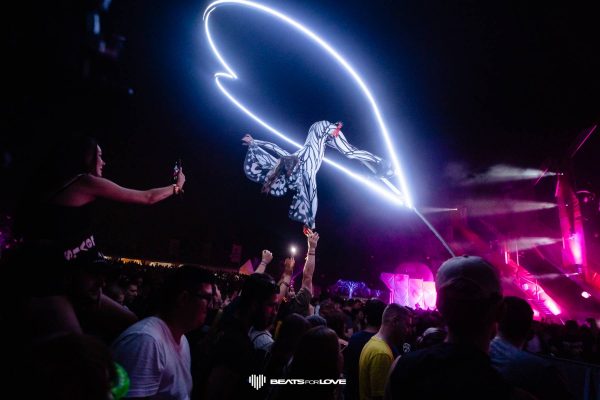 Person in a butterfly costume elevated above a crowd at a nighttime music festival with bright neon light trails.