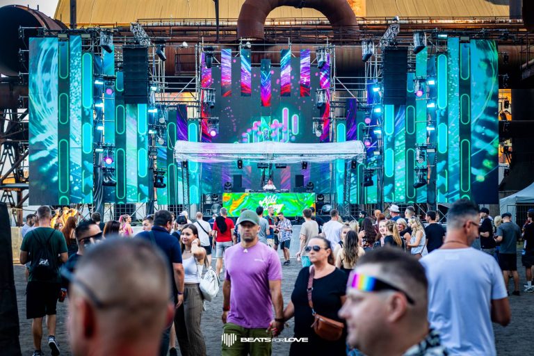 Crowd of people walking and standing in front of a large outdoor music stage with colorful LED screens and a DJ performing.