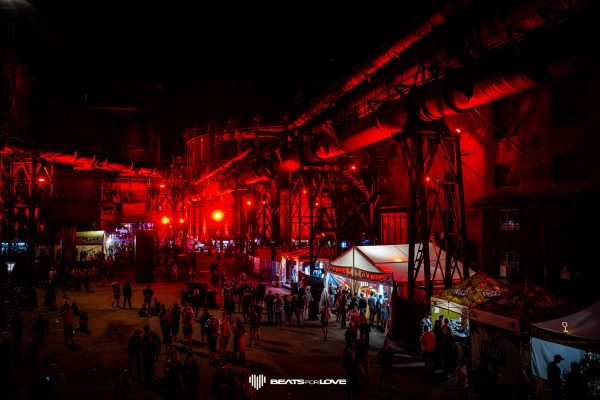 Crowd of people at night walking and gathering near red-lit industrial structures with vendor tents and stalls at a festival.