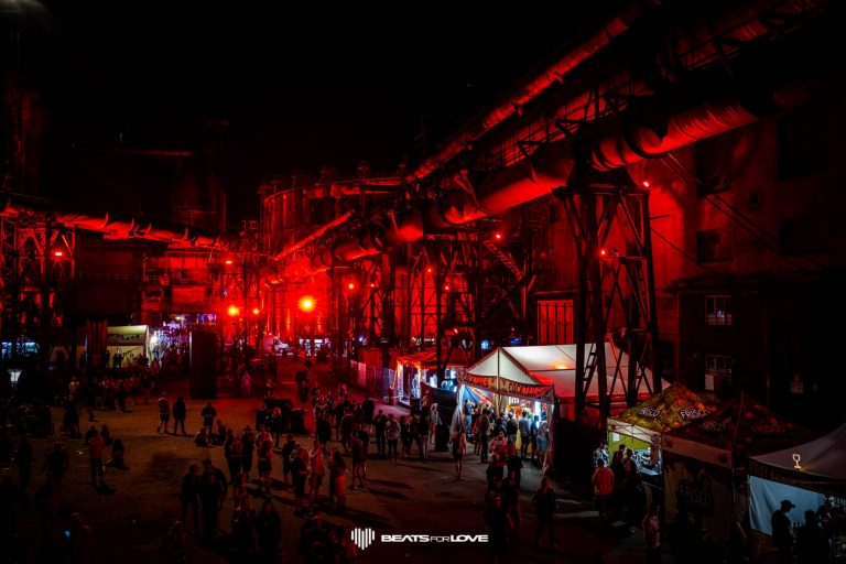 Crowd of people at night walking and gathering near red-lit industrial structures with vendor tents and stalls at a festival.