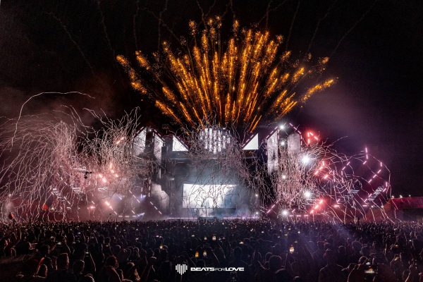 Large crowd at a nighttime music festival with fireworks and streamers lighting up the sky above the stage.
