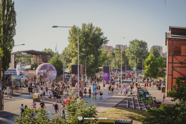 Crowd of people walking and gathering outdoors in a park-like area with trees, streetlights, and buildings in the background during a sunny day.
