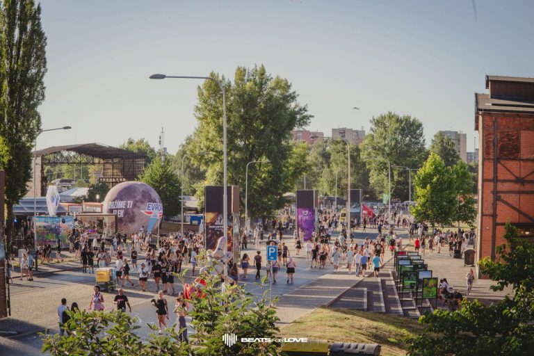 Crowd of people walking and gathering outdoors in a park-like area with trees, streetlights, and buildings in the background during a sunny day.