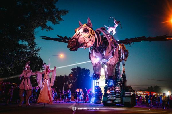 Large mechanical winged horse sculpture with a performer on top and two costumed people on stilts, surrounded by a crowd at dusk.