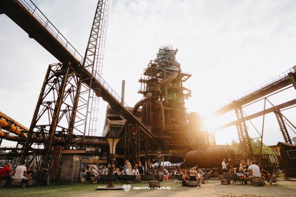 Outdoor gathering of people sitting on benches and chairs beneath large industrial metal structures at sunset with 