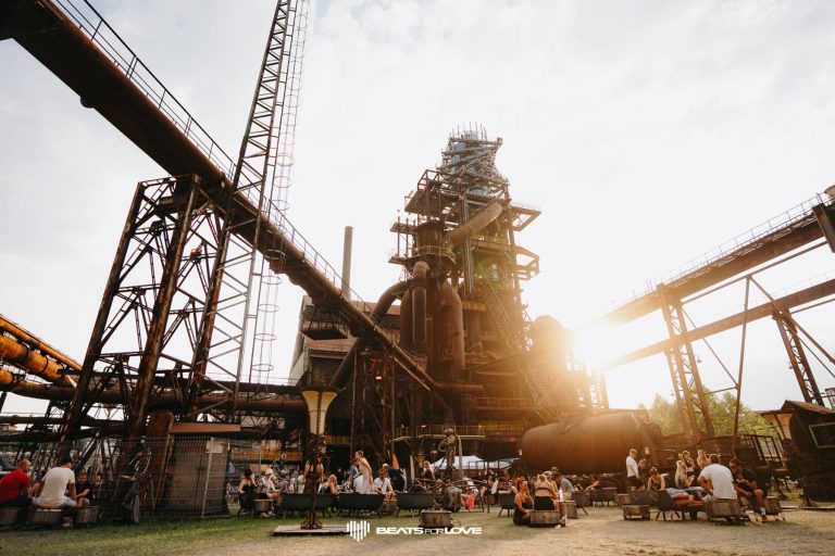Outdoor gathering of people sitting on benches and chairs beneath large industrial metal structures at sunset with 