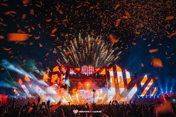 Crowd at a nighttime outdoor concert with bright orange stage lights, fireworks, confetti, and cheering audience.