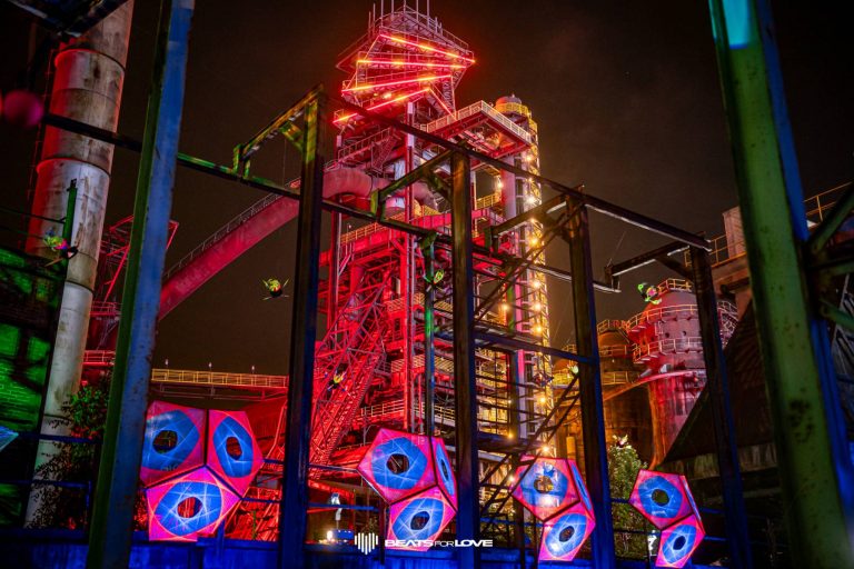 Industrial structure lit with bright red, yellow, and blue neon lights at night with colorful geometric shapes and metallic framework in the foreground.