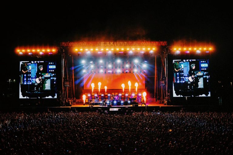 Large outdoor concert stage with bright lights, fire effects, two giant screens showing musicians, and a large crowd at night.