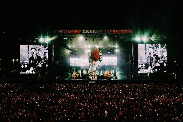 Large crowd at night watching a band on stage with bright lights and big screens showing a guitarist singing into a microphone