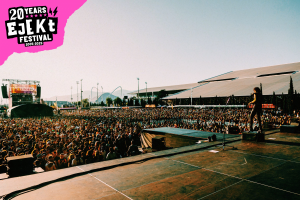 Large crowd gathered in an outdoor festival setting with a performer on stage during daylight and a 20 Years Ejekt Festival logo in the corner.
