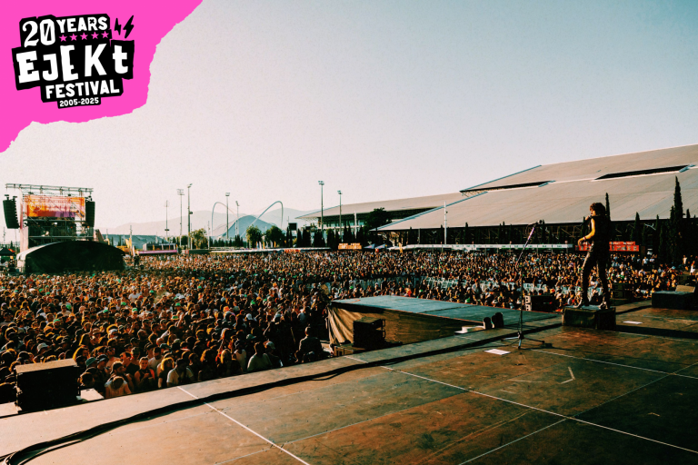 Large crowd gathered in an outdoor festival setting with a performer on stage during daylight and a 20 Years Ejekt Festival logo in the corner.
