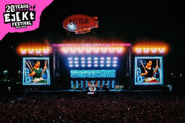 Night view of a large outdoor concert stage with bright lights, big screens showing a drummer, a band performing, a crowd, and a blimp labeled 