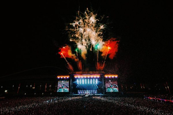 Large outdoor concert at night with a brightly lit stage, huge crowd, and colorful red, green, and white fireworks overhead