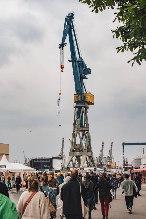 Large blue and yellow industrial crane at a busy outdoor event with many people walking nearby under a cloudy sky.