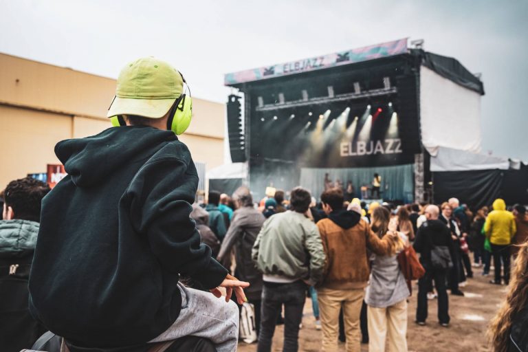 Child with green ear protection and cap facing a crowd watching a concert on an outdoor stage labeled ELBJAZZ.
