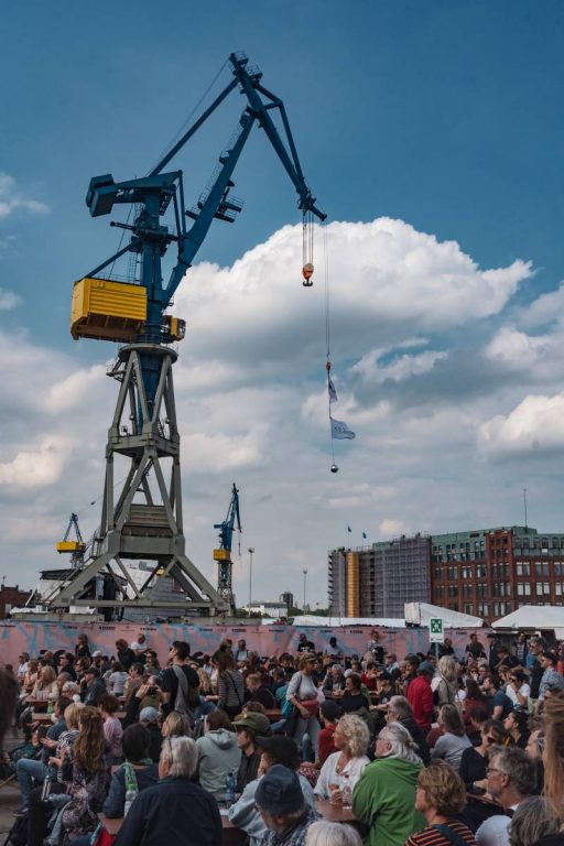 Large group of people gathered outdoors near industrial cranes and buildings under a partly cloudy sky.