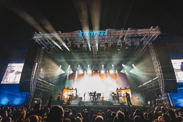 Outdoor night concert stage with bright lights and silhouettes of musicians performing in front of a crowd.
