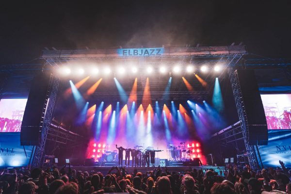 Band members taking a bow on stage with colorful stage lights and a large crowd at the Elbjazz music festival.