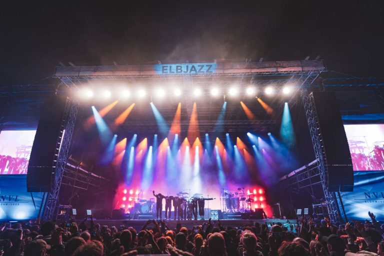 Band members taking a bow on stage with colorful stage lights and a large crowd at the Elbjazz music festival.