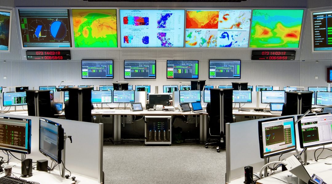 Control room filled with multiple computer monitors displaying data and maps, large colorful weather and system analysis screens on the wall above.