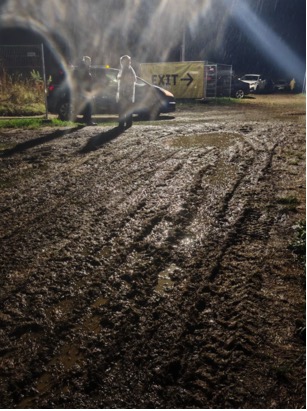 Two people standing near cars on a muddy ground at night with a large illuminated "EXIT" sign in the background pointing right.