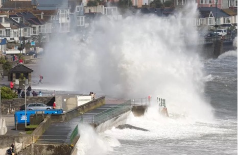 Large ocean waves crashing over a seaside town's walkway and buildings during rough sea conditions.