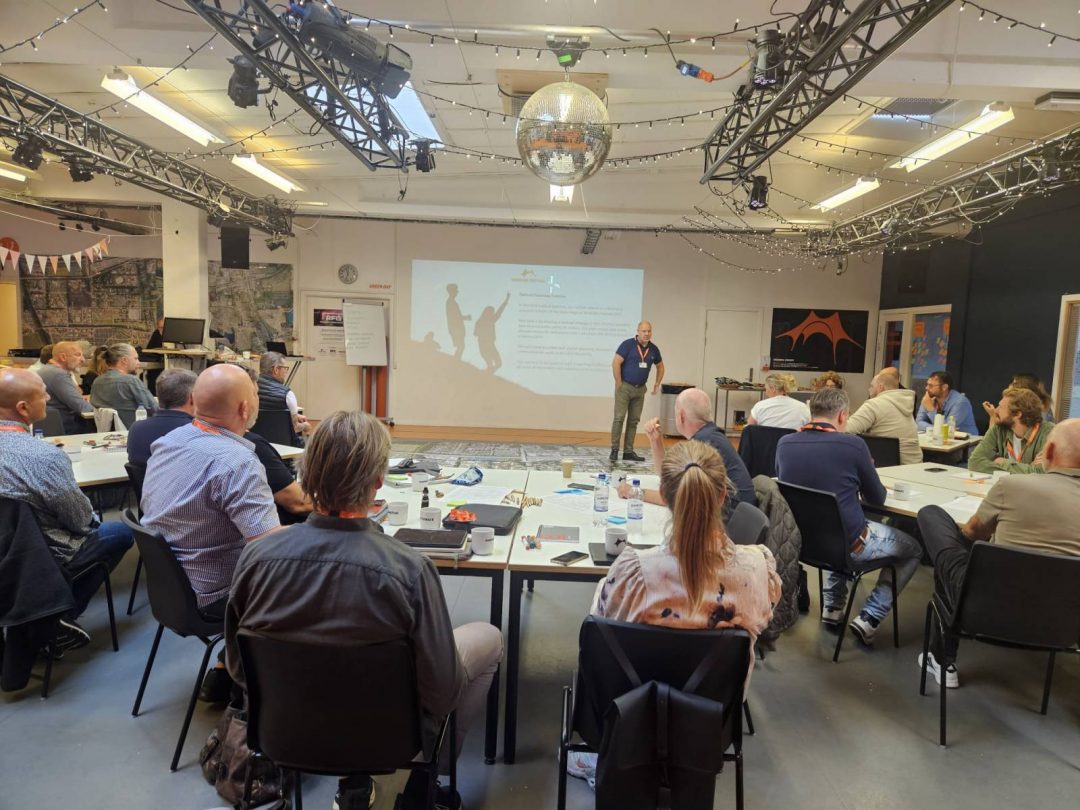 Group of people sitting at tables attentively listening to a presenter standing near a projection screen in a decorated indoor conference room.