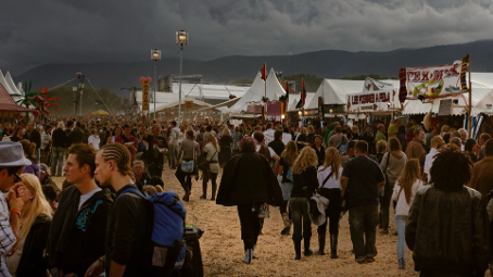 Crowd of people walking between food and merchandise tents at an outdoor festival under a dark cloudy sky.