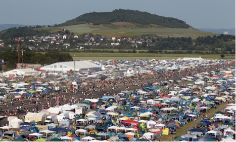 Large outdoor event with numerous colorful tents and crowds of people, set against a backdrop of fields and a hill with scattered houses.