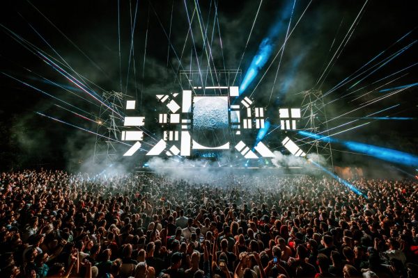 Large crowd at a nighttime concert with bright stage lights, laser beams, and a moon image on the screen.