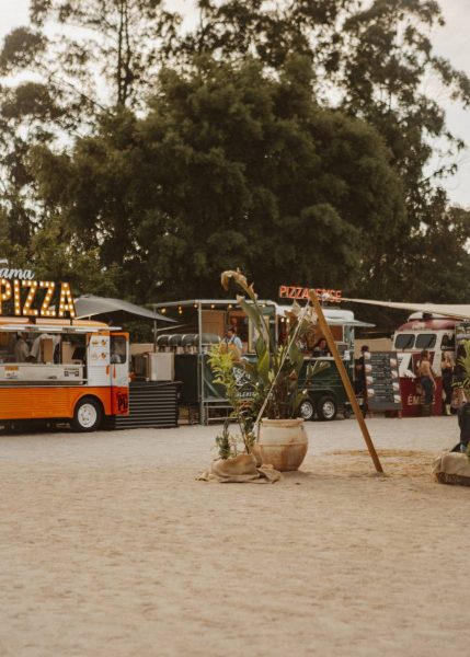 Outdoor food truck area with pizza trucks, potted plants, and people standing near the trucks under tall trees.