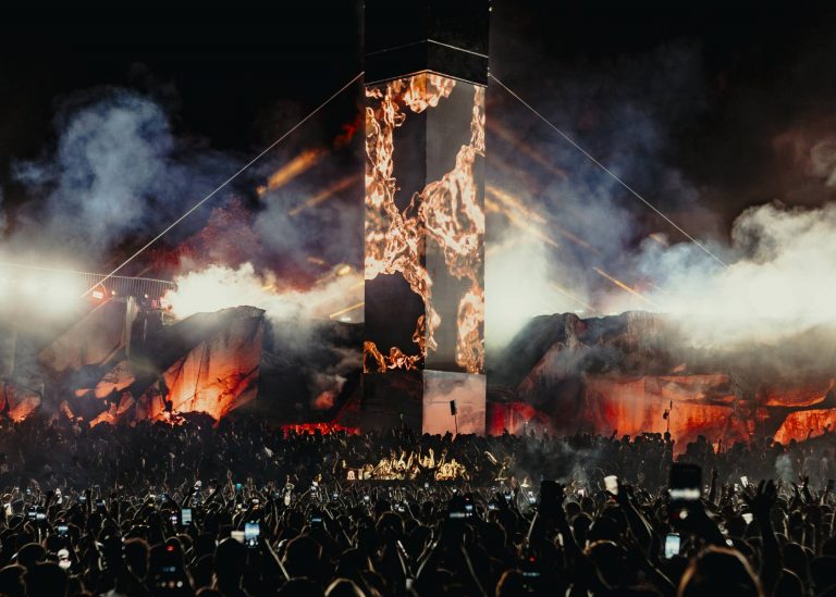 A large crowd at a nighttime outdoor concert with a tall smokey stage pillar displaying fire visuals and people holding up phones.