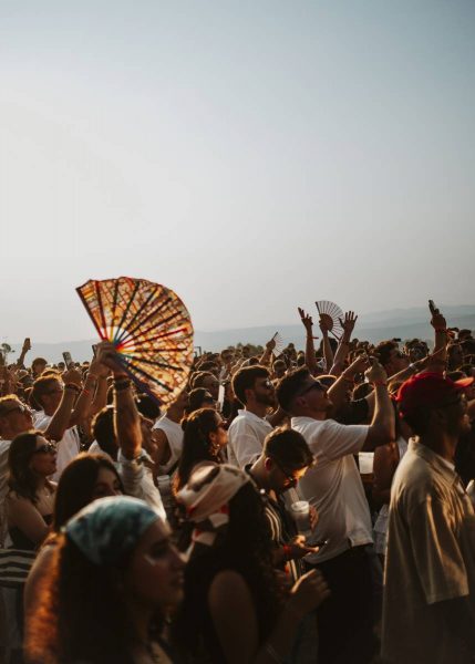 Crowd of people at an outdoor event or festival in the late afternoon, some holding handheld fans and drinks.