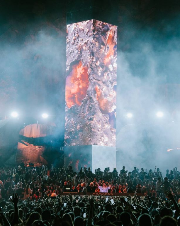 Large crowd watching a tall, illuminated rectangular structure with fiery volcanic imagery in a smoky night setting.