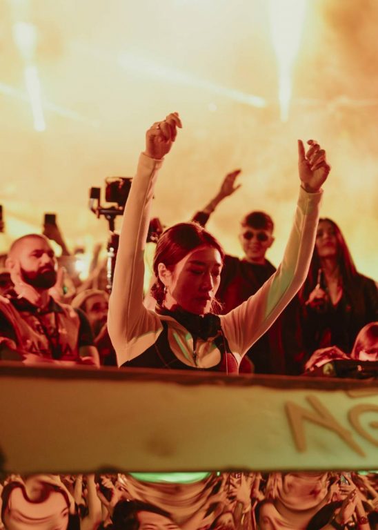 Woman DJ with arms raised performing at a lively concert with an enthusiastic crowd and bright stage lights.