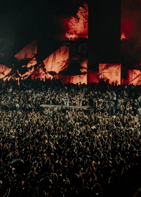 Large crowd at a nighttime outdoor event with red-lit rock formations or structures in the background.