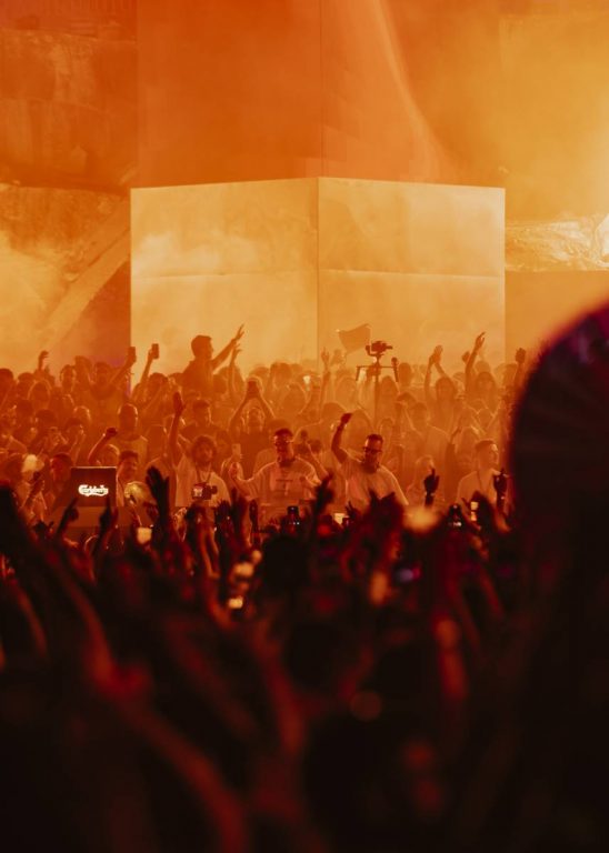 Crowd at a lively concert or event with raised hands and orange stage lighting, creating an energetic atmosphere.