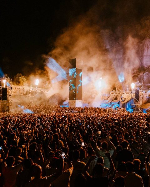 Large crowd at a nighttime outdoor concert or festival with illuminated smoke, stage lights, and a central tall screen displaying blue graphics.