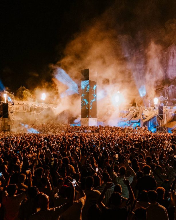 Large crowd at a nighttime outdoor concert or festival with illuminated smoke, stage lights, and a central tall screen displaying blue graphics.